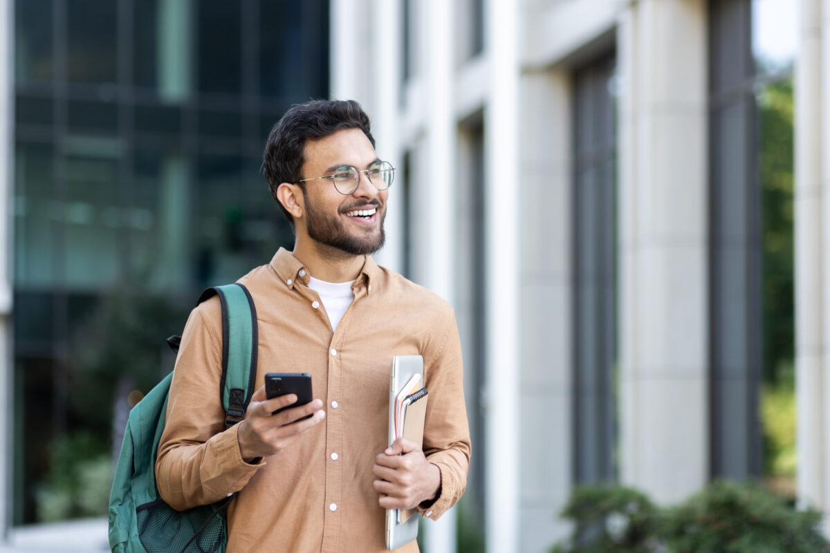 Young man walking through the city with a backpack, student smiling outside university campus in casual clothes